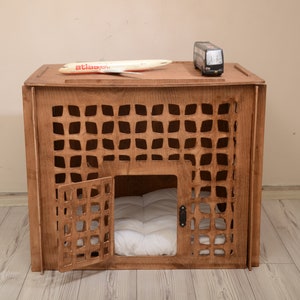 K&ouml;nnte beinhalten: Eine braune Haustierbox aus Holz mit einem gitterartigen Ausschnittmuster an der Vorder- und den Seitenw&auml;nden. Die Box verf&uuml;gt &uuml;ber eine Fl&uuml;gelt&uuml;r an der Vorderseite und ein wei&szlig;es, gestepptes Kissen im Inneren. Ein Spielzeugflugzeug und ein Bus stehen oben.