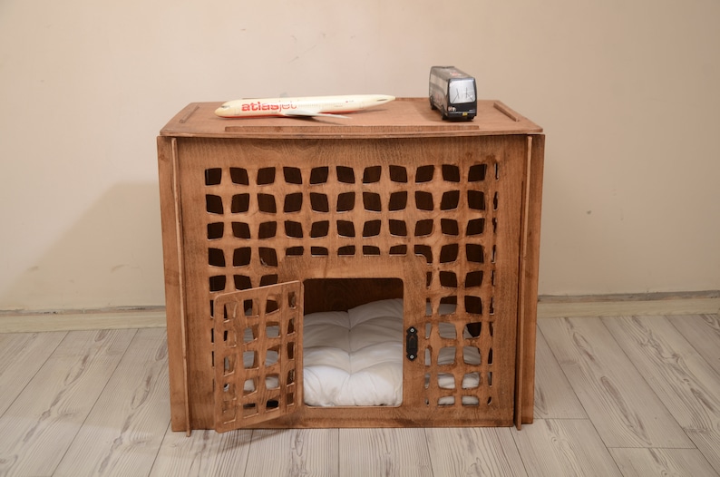K&ouml;nnte beinhalten: Eine braune Haustierbox aus Holz mit einem gitterartigen Ausschnittmuster an der Vorder- und den Seitenw&auml;nden. Die Box verf&uuml;gt &uuml;ber eine Fl&uuml;gelt&uuml;r an der Vorderseite und ein wei&szlig;es, gestepptes Kissen im Inneren. Ein Spielzeugflugzeug und ein Bus stehen oben.