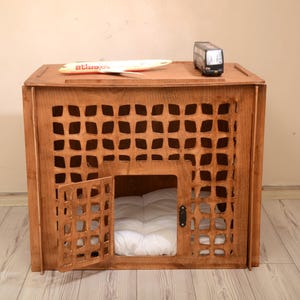 K&ouml;nnte beinhalten: Eine braune Tierbox aus Holz mit einem gitterartigen Ausschnittmuster auf der Vorderseite und einer Fl&uuml;gelt&uuml;r. Im Inneren der Box liegt ein wei&szlig;es, gestepptes Kissen. Ein kleines Spielzeugflugzeug und ein Spielzeugbus stehen auf der Oberseite.