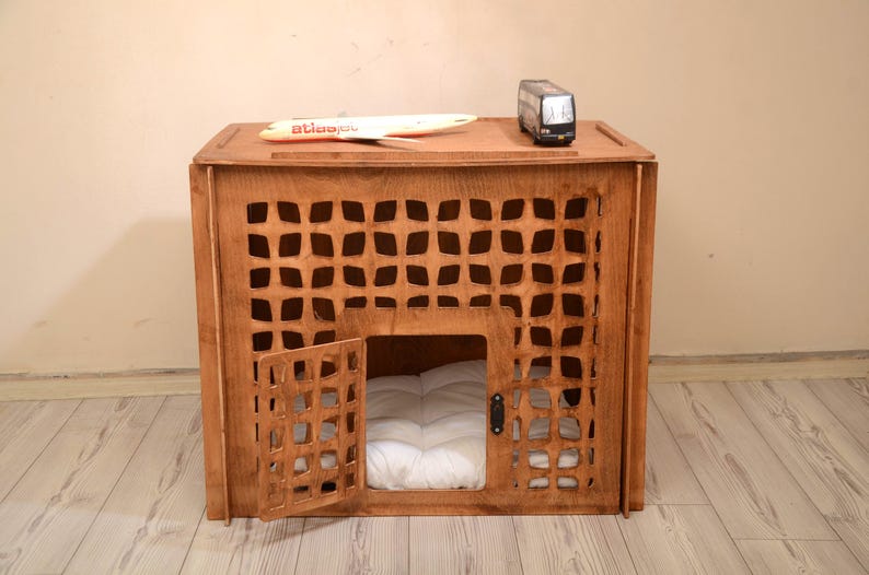 K&ouml;nnte beinhalten: Eine braune Tierbox aus Holz mit einem gitterartigen Ausschnittmuster auf der Vorderseite und einer Fl&uuml;gelt&uuml;r. Im Inneren der Box liegt ein wei&szlig;es, gestepptes Kissen. Ein kleines Spielzeugflugzeug und ein Spielzeugbus stehen auf der Oberseite.