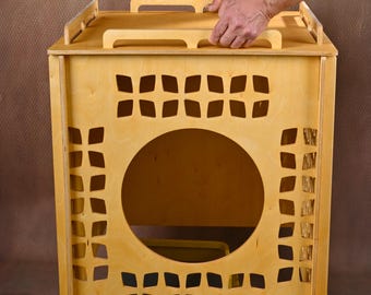 May include: A light-colored wooden cat house with a square shape and decorative cutouts. The front features a large circular opening and a removable tray. The top has two handles for easy carrying. The cat house is placed over a gray and yellow litter box.