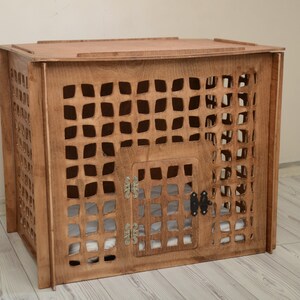 K&ouml;nnte beinhalten: Eine rechteckige Haustierbox aus Holz mit mittelbraunem Finish und einem gitterartigen Ausschnittmuster an allen Seiten. Die Vorderseite verf&uuml;gt &uuml;ber eine Fl&uuml;gelt&uuml;r mit silberfarbenen Metallscharnieren und einem schwarzen Riegel.