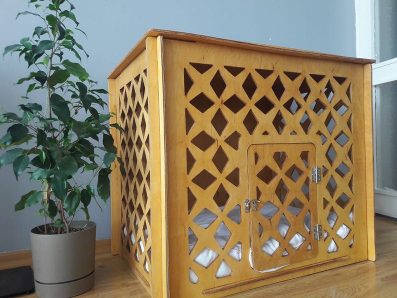 May include: A light-colored wooden pet crate featuring a decorative lattice pattern on the sides and front. The structure includes a hinged door with a metal latch and a white cushion inside, providing a contained space for a small animal.