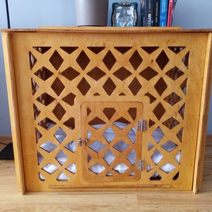 May include: A light brown wooden pet crate featuring a decorative lattice pattern across the front panel. The structure includes a central hinged door with a metal latch and hinges, and a soft white cushion is visible inside the enclosure.
