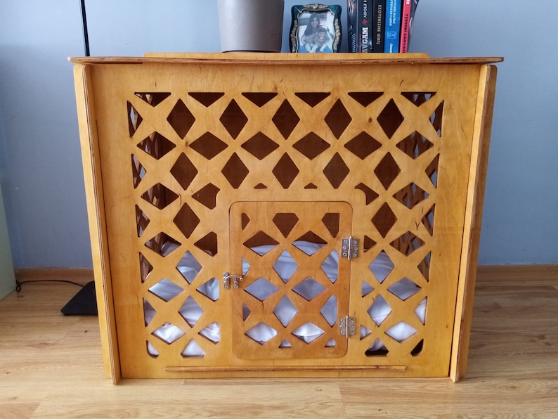 May include: A light brown wooden pet crate featuring a decorative lattice pattern across the front panel. The structure includes a central hinged door with a metal latch and hinges, and a soft white cushion is visible inside the enclosure.
