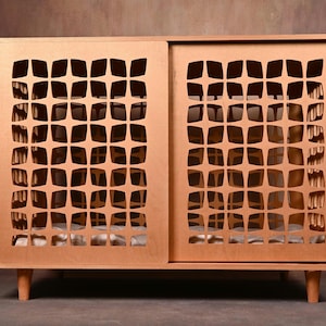 Könnte beinhalten: Eine rechteckige Haustierbox aus Holz in hellbrauner Ausführung mit zwei Schiebetüren, die ein sich wiederholendes geometrisches Sternmuster aufweisen. Die Konstruktion steht auf vier kurzen, konisch zulaufenden Beinen und hat ein Kissen.