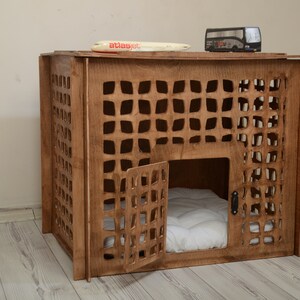 K&ouml;nnte beinhalten: Eine braune Haustierbox aus Holz mit einem gitterartigen Ausschnittmuster an den Seiten und der Vordert&uuml;r. Im Inneren befindet sich ein wei&szlig;es, gestepptes Kissen. Ein kleines Spielzeugflugzeug mit der Aufschrift atlasjet und ein Spielzeugbus stehen oben auf.