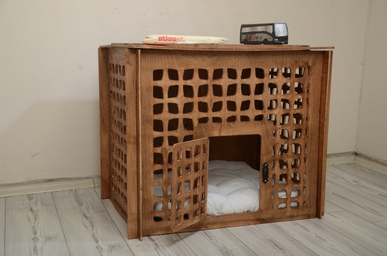 K&ouml;nnte beinhalten: Eine braune Haustierbox aus Holz mit einem gitterartigen Ausschnittmuster an den Seiten und der Vordert&uuml;r. Im Inneren befindet sich ein wei&szlig;es, gestepptes Kissen. Ein kleines Spielzeugflugzeug mit der Aufschrift atlasjet und ein Spielzeugbus stehen oben auf.