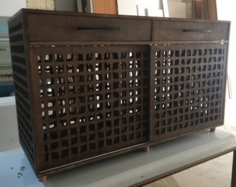 May include: A dark brown wooden cabinet with two drawers and a sliding door. The door features a geometric cut-out design. The cabinet has black handles and sits on small wooden feet. The overall design is modern and minimalist.