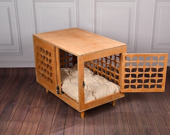 May include: A rectangular wooden pet crate with a light brown finish, featuring a geometric cutout pattern on the side and front door panels. The crate is elevated on four short legs and contains a tufted, cream-colored rectangular cushion inside.