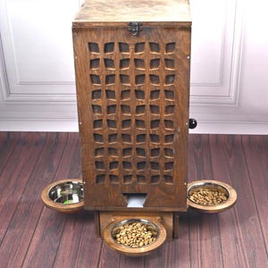 Puede incluir: Un comedero de madera para mascotas con acabado marrón oscuro. El comedero tiene un diseño rectangular con un patrón decorativo recortado en la parte delantera. Cuenta con tres cuencos de acero inoxidable para comida y agua.