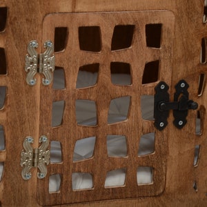 K&ouml;nnte beinhalten: Eine braune Holzkiste mit einem gitterartigen Ausschnittmuster und einer Fl&uuml;gelt&uuml;r. Die T&uuml;r ist mit zwei verzierten silberfarbenen Metallscharnieren befestigt und mit einem schwarzen Metallriegel gesichert. Das Innere enth&auml;lt ein wei&szlig;es Stofffutter.