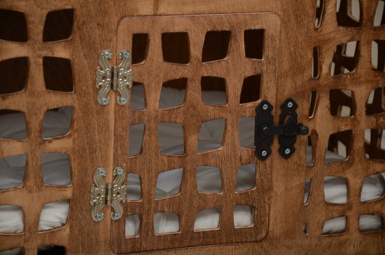 K&ouml;nnte beinhalten: Eine braune Holzkiste mit einem gitterartigen Ausschnittmuster und einer Fl&uuml;gelt&uuml;r. Die T&uuml;r ist mit zwei verzierten silberfarbenen Metallscharnieren befestigt und mit einem schwarzen Metallriegel gesichert. Das Innere enth&auml;lt ein wei&szlig;es Stofffutter.