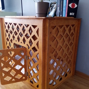 May include: A light brown wooden pet crate featuring a decorative lattice pattern on the side panels and a hinged door. The structure has a flat top surface and contains a white fabric cushion inside, providing a contained space for a pet.