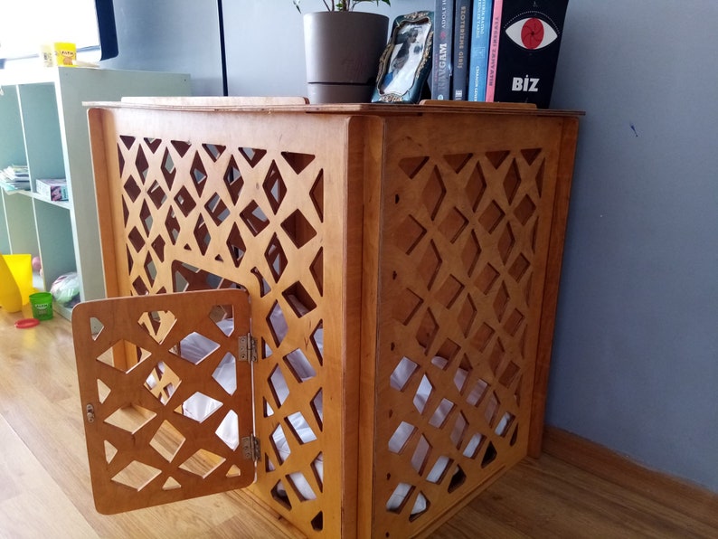 May include: A light brown wooden pet crate featuring a decorative lattice pattern on the side panels and a hinged door. The structure has a flat top surface and contains a white fabric cushion inside, providing a contained space for a pet.