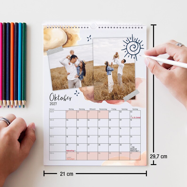May include: A wall calendar for October 2027 featuring two photographs of a family in a field, watercolor accents, and a hand-drawn sun. The calendar measures 8.27 inches by 11.69 inches, indicated by black dimension lines and text.