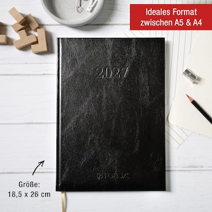 Könnte beinhalten: Ein schwarzer 2027 Planer mit texturiertem Einband und dem unten eingeprägten Markennamen TOBJA. Der Planer misst 18,5 cm x 26 cm. Er verfügt über ein Lesebändchen und wird zusammen mit einem Bleistift, einem Spitzer und Holzklötzen gezeigt.