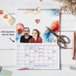 May include: A 2027 August wall calendar featuring a watercolor background with blue and orange splashes, a cutout portrait of a couple, and a red heart sticker. The calendar grid includes handwritten notes, and the page is displayed with scissors.