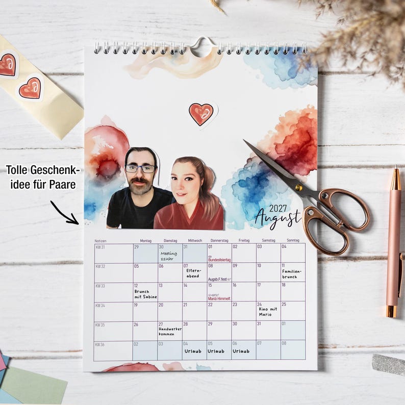 May include: A 2027 August wall calendar featuring a watercolor background with blue and orange splashes, a cutout portrait of a couple, and a red heart sticker. The calendar grid includes handwritten notes, and the page is displayed with scissors.