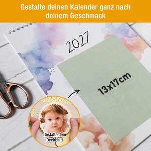 May include: A 2027 wall calendar with watercolor accents and a sage green rectangular overlay labeled 13x17cm. A circular inset shows a scalloped-edge photo of a baby with the German text Gestalte dein Deckblatt. Copper scissors are visible.