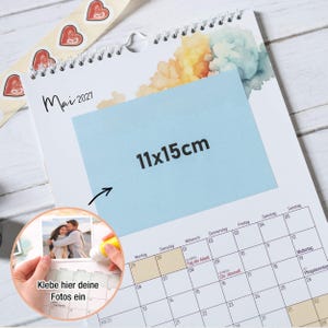 May include: A wall calendar for May 2027 featuring a spiral binding at the top and a light blue rectangular overlay with the text 11x15cm. A circular inset shows a person applying a photograph to the calendar with the text Klebe hier deine Fotos ein.