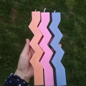 May include: Three tall candles with a zig-zag pattern. The candles are peach, pink, and light blue.