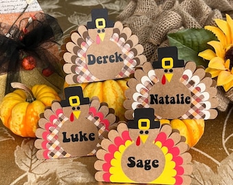 May include: Four Thanksgiving turkey place card holders with the names Derek, Natalie, Luke, and Sage. Each turkey is made of brown cardstock with a plaid pattern and a black hat. The turkeys are sitting on a brown table with a burlap background.
