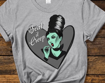 May include: Gray t-shirt with a graphic of a woman with green skin and black hair in a bouffant hairstyle. The woman is holding a lollipop and the text "Beat it Creep" is written in a heart shape around her.