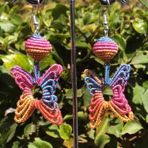May include: A pair of colorful butterfly earrings with a pink, yellow, and blue color scheme. The earrings are made of string and have a small ball at the top.