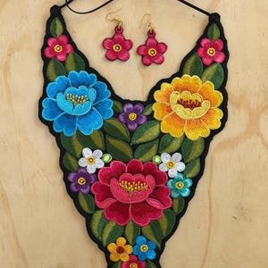 May include: Embroidered floral necklace and matching earrings. The necklace features a black base with colorful embroidered flowers in shades of blue, yellow, red, and pink. The earrings are small, pink flower shapes.