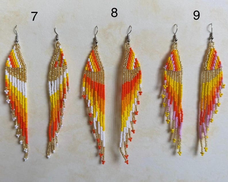 May include: Handmade beaded earrings in a feather design. The earrings feature a gold top with a gradient of colors including orange, yellow, white, and red. The earrings have a fringe of beads at the bottom.