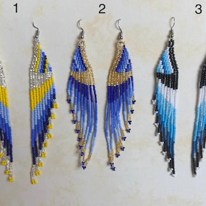 May include: Three pairs of beaded dangle earrings. The earrings feature intricate patterns in blue, yellow, silver, black, and white. Each pair has a unique design, with variations in color and bead arrangement. The earrings have silver-tone hooks.