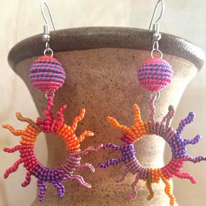 May include: A pair of colourful crocheted earrings with a sunburst design. The earrings are made of pink, orange, yellow, green, and purple yarn. Each earring has a small pink and blue sphere hanging from it.