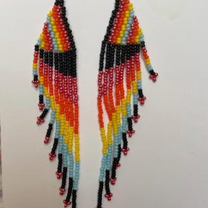 May include: A pair of beaded earrings with a colorful design. The earrings feature a triangular top with a rainbow pattern of red, orange, yellow, blue, and black beads. Long black beaded fringe hangs from the bottom, with red accents.