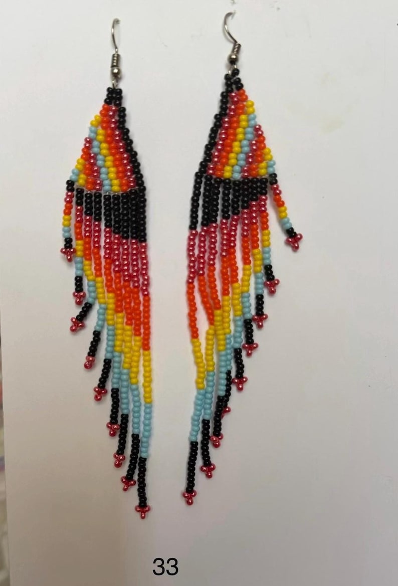 May include: A pair of beaded earrings with a colorful design. The earrings feature a triangular top with a rainbow pattern of red, orange, yellow, blue, and black beads. Long black beaded fringe hangs from the bottom, with red accents.