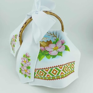 May include: A white fabric-lined basket with a woven handle, decorated with embroidered floral and geometric patterns. The embroidery includes pink flowers, green leaves, and a colourful geometric border. A white cloth is tied to the handle.