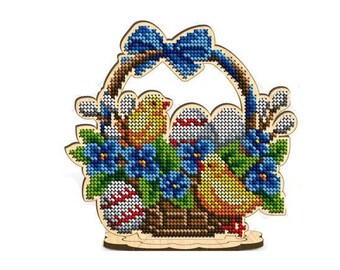Può includere: Un cestino di Pasqua in legno con un fiocco blu, pieno di uova colorate, fiori e due uccellini gialli. Il cestino è decorato con un motivo floreale e ha un piedistallo in legno.