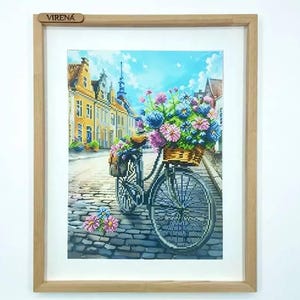 May include: A framed cross-stitch embroidery depicting a bicycle on a cobblestone street in a European town. The bicycle features a wicker basket filled with pink and blue flowers. The word VIRENA is printed on the top of the wooden frame.