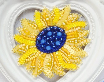 May include: A yellow and blue beaded sunflower brooch in a white heart-shaped frame. The brooch is surrounded by yellow and blue beads.