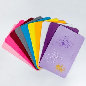 Könnte beinhalten: Ein Set aus zehn Filzblättern in verschiedenen Farben, darunter rosa, rot, grau, blau, gelb, weiß und lila. Die Blätter sind fächerförmig angeordnet, wobei das lila Blatt oben liegt. Das lila Blatt ist mit verschiedenen kleinen Perlen und Pailletten beklebt, die ein dekoratives Muster bilden.