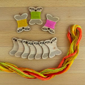 Könnte beinhalten: Holzspulen für Stickgarn mit farbigem Garn. Drei Spulen sind mit gelbem, grünem und rosa Garn gewickelt. Sieben weitere Spulen sind leer. Ein Strang mehrfarbigen Garns in Orange, Gelb und Grün ist ebenfalls vorhanden.