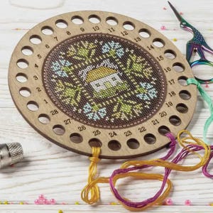 Könnte beinhalten: Ein hölzerner Stickgarn-Organizer mit einem Kreuzstich-Design mit einem Haus und floralen Mustern. Der Organizer hat mehrere Löcher für den Faden und wird von bunten Fäden und einer Schere begleitet. Das Design ist in Grün-, Blau- und Gelbtönen gehalten.