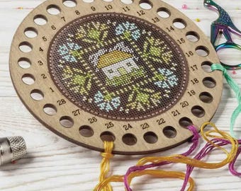 May include: A wooden embroidery floss organizer with a cross-stitch design featuring a house and floral pattern. The organizer has multiple holes for thread and is accompanied by colorful threads and scissors. The design is in shades of green, blue, and yellow.