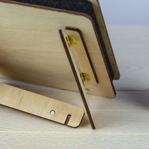 May include: A wooden picture frame with a black backing and gold hinges. The frame is standing on a light wood surface.