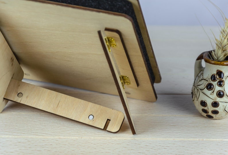May include: A wooden picture frame with a black backing and gold hinges. The frame is standing on a light wood surface.
