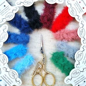 May include: Assortment of colorful faux fur trim, each piece is 16 meters long and made of 100% nylon. The trim is arranged in a circle around a pair of gold scissors. The text on the labels reads "Tela Artis" and "100% нейлон, 16м".