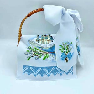 May include: White embroidered fabric basket liner featuring a dove, a church, and a bouquet of flowers. The liner has a blue and white geometric border.