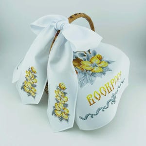 May include: A white Easter basket cover and bow with embroidered yellow flowers and the word "Воскресение" in gold and grey. The basket has a woven handle.