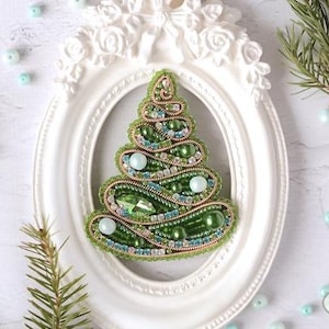 Könnte beinhalten: Eine grüne, perlenbesetzte Weihnachtsbaum-Brosche mit goldenen und weißen Akzenten. Die Brosche wird in einem weißen ovalen Rahmen präsentiert.