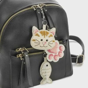 May include: A black backpack with a cat-themed keyring. The keyring includes a stitched cat face, a pink paw print, and a fish. The cat face is white with orange stripes and black whiskers. The paw print is pink and white.